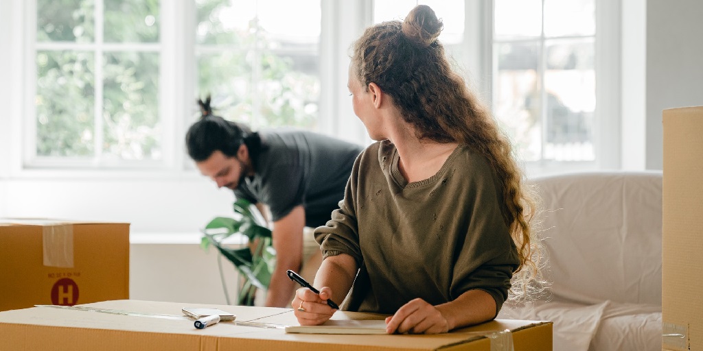 One person writing packing labels on a moving box looks back at another person who is bent down doing something, beside a piece of furniture covered in a white sheet