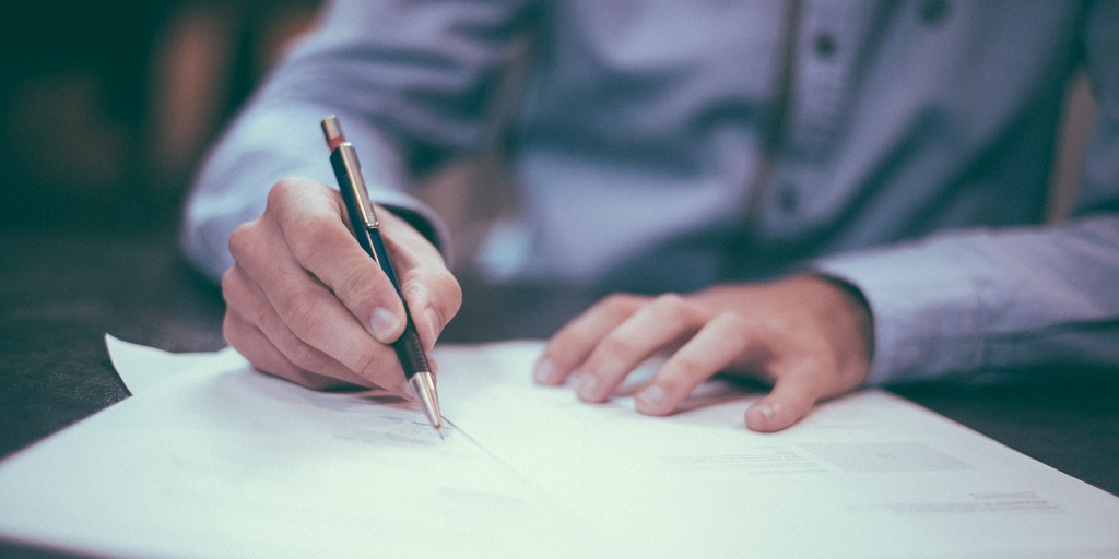 Person holding pen with hands resting on papers on a flat surface infront of them