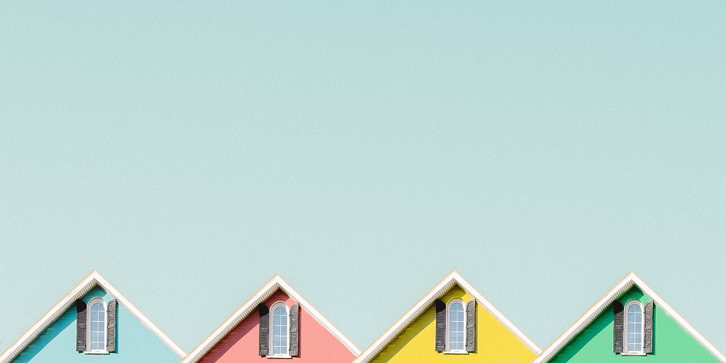 Peaks of four beach huts (one blue, pink, yellow, green), each with a window in the peak of the roof, against cloudless blue sky