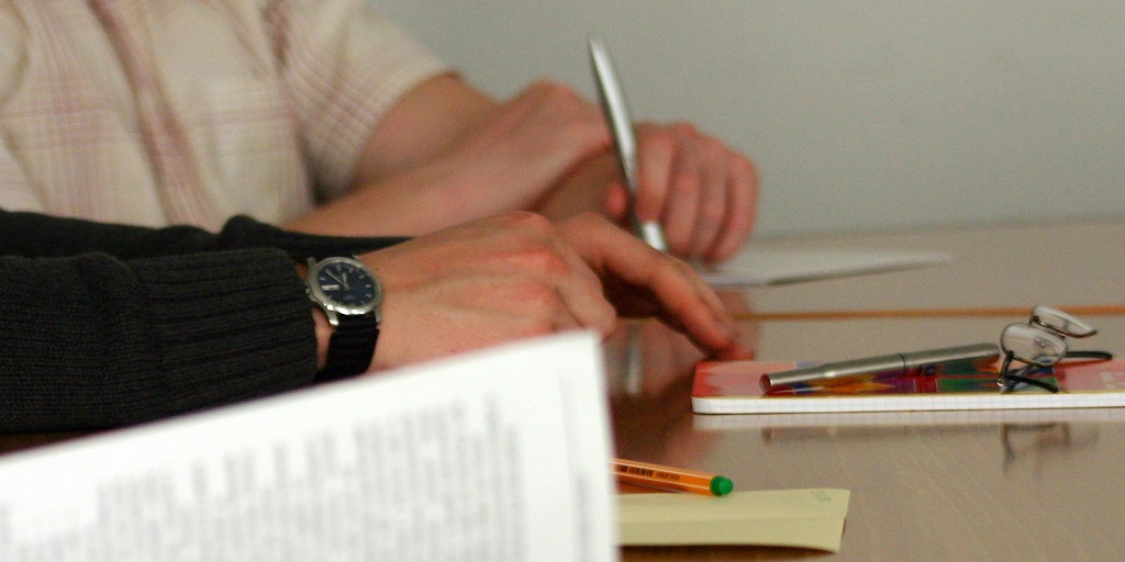 Two people sat at a desk, one holding a pen, there are papers and another pen on the desk and some glasses