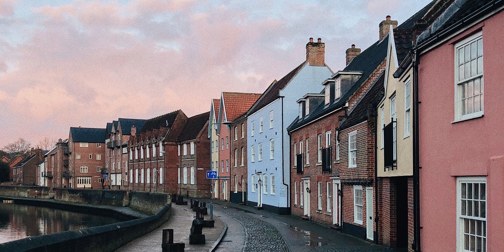 Houses facing a river, with a cobbled road infront of the houses. Pink clouds in the sky
