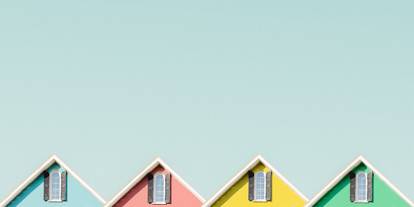 Peaks of four beach huts (one blue, pink, yellow, green), each with a window in the peak of the roof, against cloudless blue sky